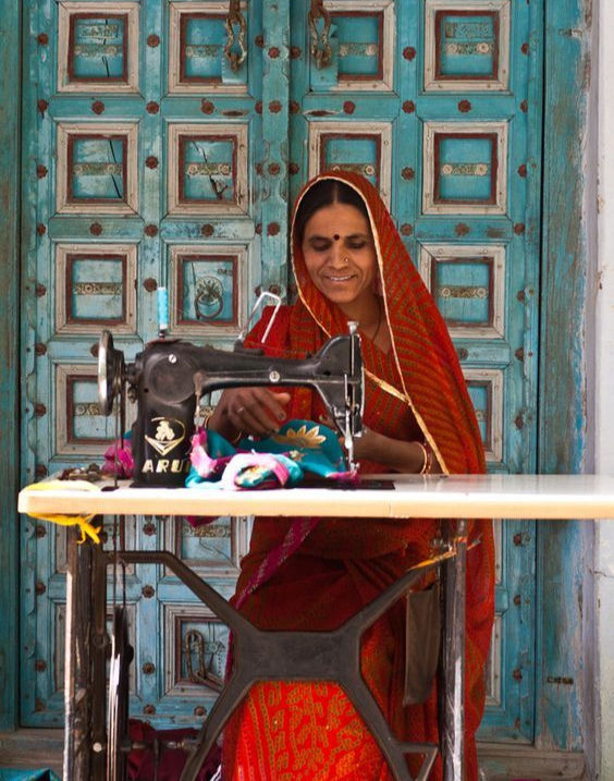 A woman in a vibrant red saree skillfully sewing on a manual sewing machine, set against a beautifully carved blue wooden door in a rural setting.
