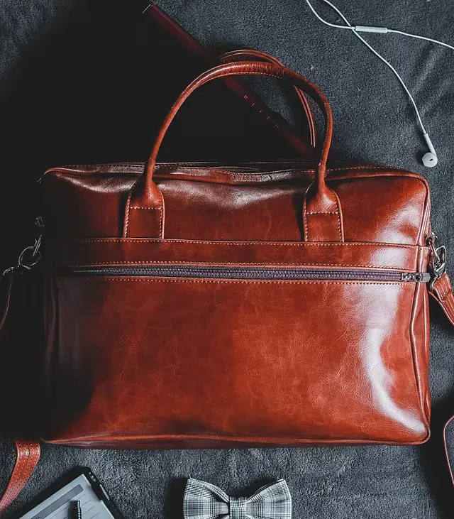 Brown leather bag on a dark fabric surface, surrounded by a grey plaid bow tie and white earphones. Stylish and elegant setup.