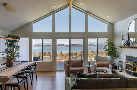 Wide view of an open-concept living and dining area with vaulted ceilings. Floor-to-ceiling windows overlook the water, featuring a wood dining table with black chairs and a mix of green and terracotta upholstered seating.