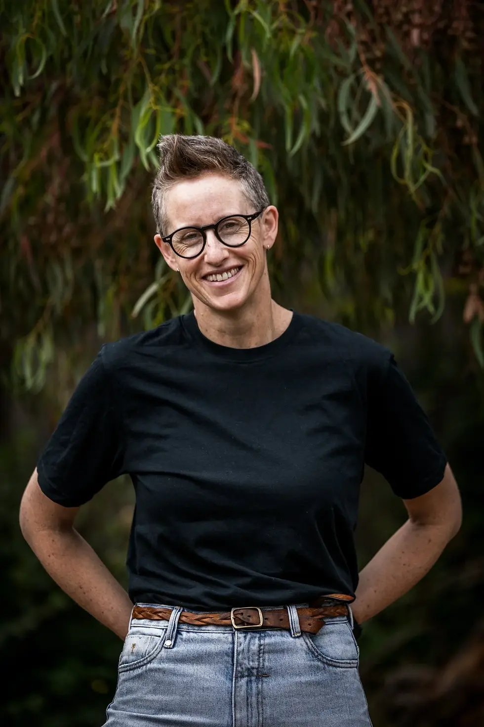 Nat Mendham, a woman in a black tshirt and jeans, smiles at the camera. She stands in front of a tree.