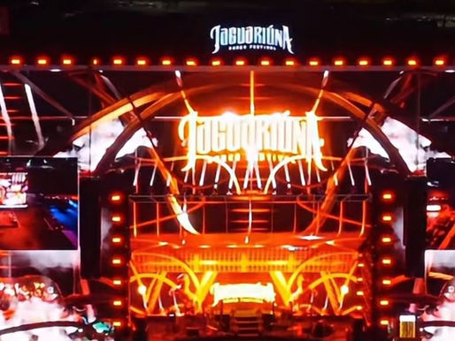 Palco de show com luzes laranja e vermelhas, telões e logo Jaguariúna, criando clima vibrante.