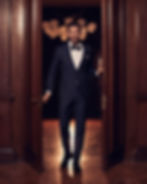 A sophisticated man in a tailored navy tuxedo with a black bow tie steps confidently through a grand wooden doorway, with warm, ambient lighting and an elegant chandelier in the background.