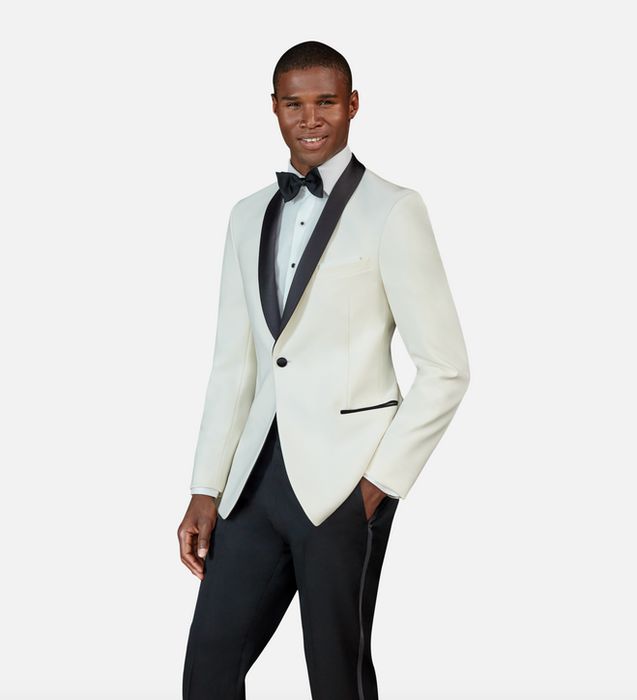 A man wearing a white dinner jacket with a black satin shawl lapel, black dress pants, a white dress shirt, black bow tie, and black buttons, posing confidently against a light grey background.