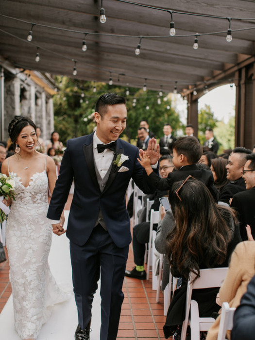 A smiling Asian groom in a navy tuxedo and bride in a white lace gown walking hand-in-hand down the aisle while guests cheer under string lights.