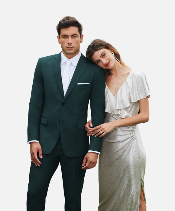 A man wearing a notch lapel green suit with a white dress shirt, white tie, and white pocket square standing confidently beside a woman in a shimmering silver gown with a ruffled neckline and high slit, resting her head on his shoulder, both posing elegantly against a light grey background.