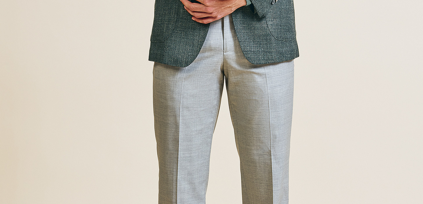 A man wearing a tailored dark sage green blazer over a light soft green turtleneck, paired with light gray trousers and brown suede lace-up shoes, completed with a silk pocket square for a polished, sophisticated look.
