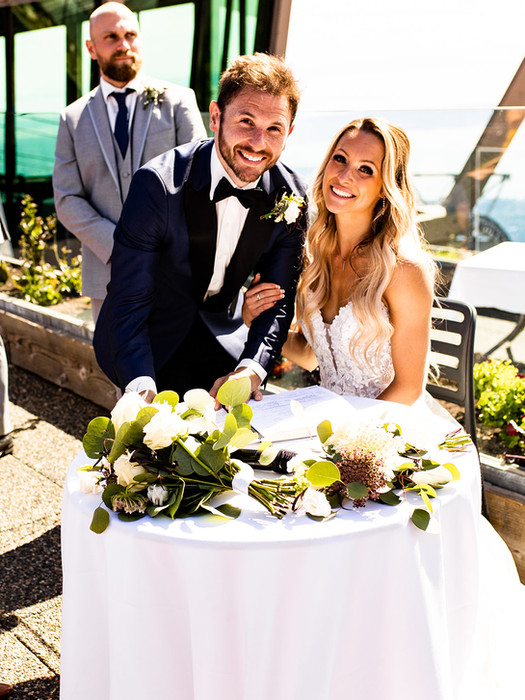 A smiling Caucasian groom in a navy tuxedo and bride in a white wedding gown posing at a decorated table adorned with flowers and greenery on a sunny day.