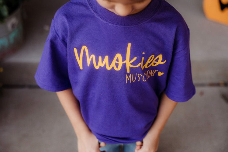 Thumbnail: Smiling girl posing in Muscatine Muskies youth tee – Letters from Elliott