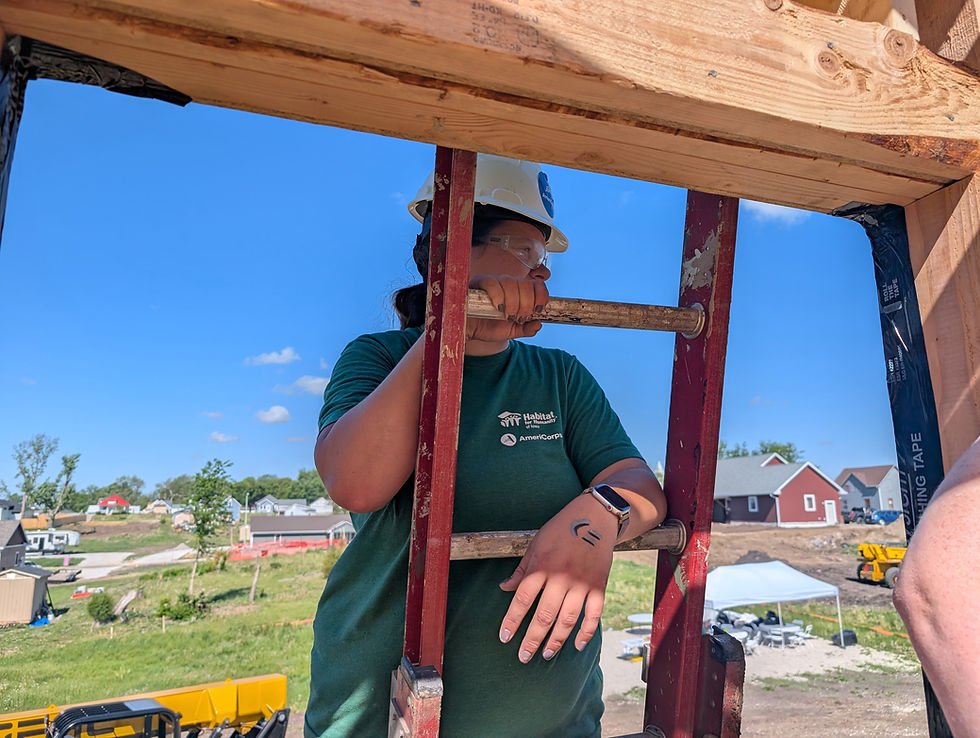 A person in a hard hat and green shirt on a ladder at a construction site. The shirt reads "Habitat for Humanity AmeriCorps." Sunny day.