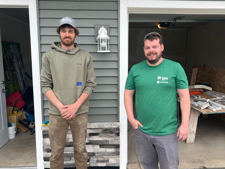 1 man with hands clasped in the middle standing next to another man in a bright green shirt. Both are standing on a driveway in front of a house.