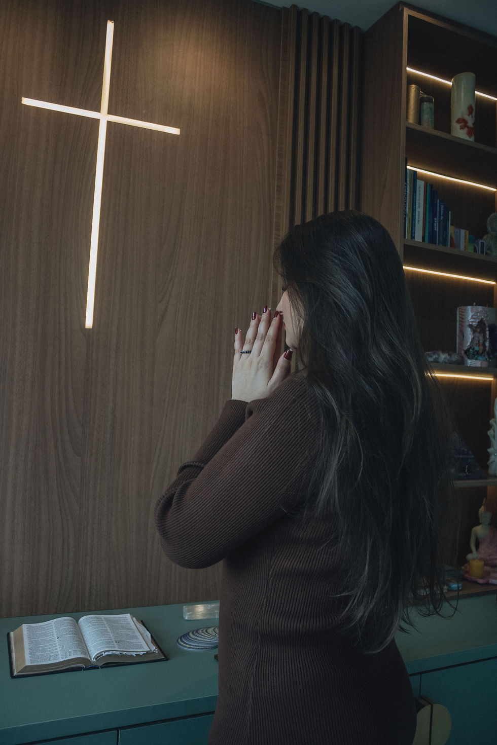 Woman with hands clasped prays before a lit cross in a wood-paneled room, with an open Bible and shelves behind her.