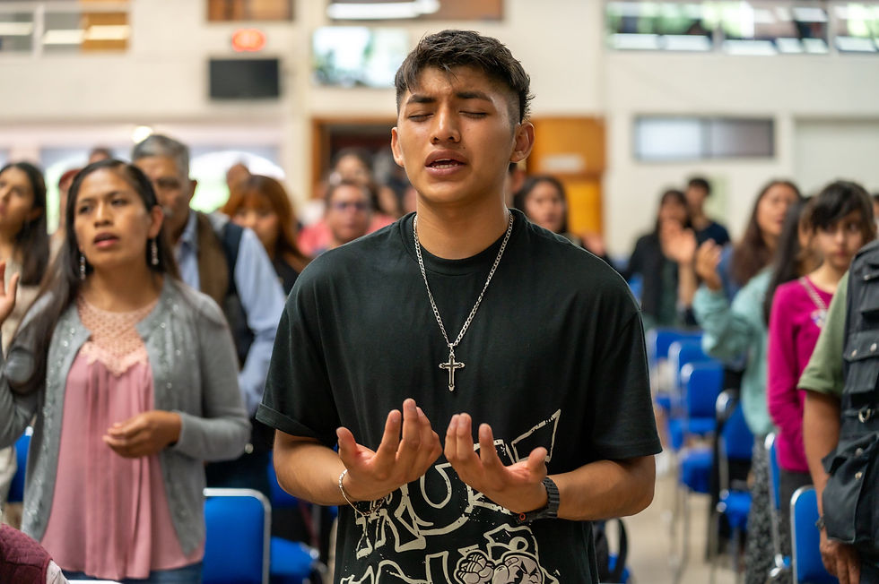 Young man with cross necklace prays with eyes closed in a church hall, congregation blurred behind him.