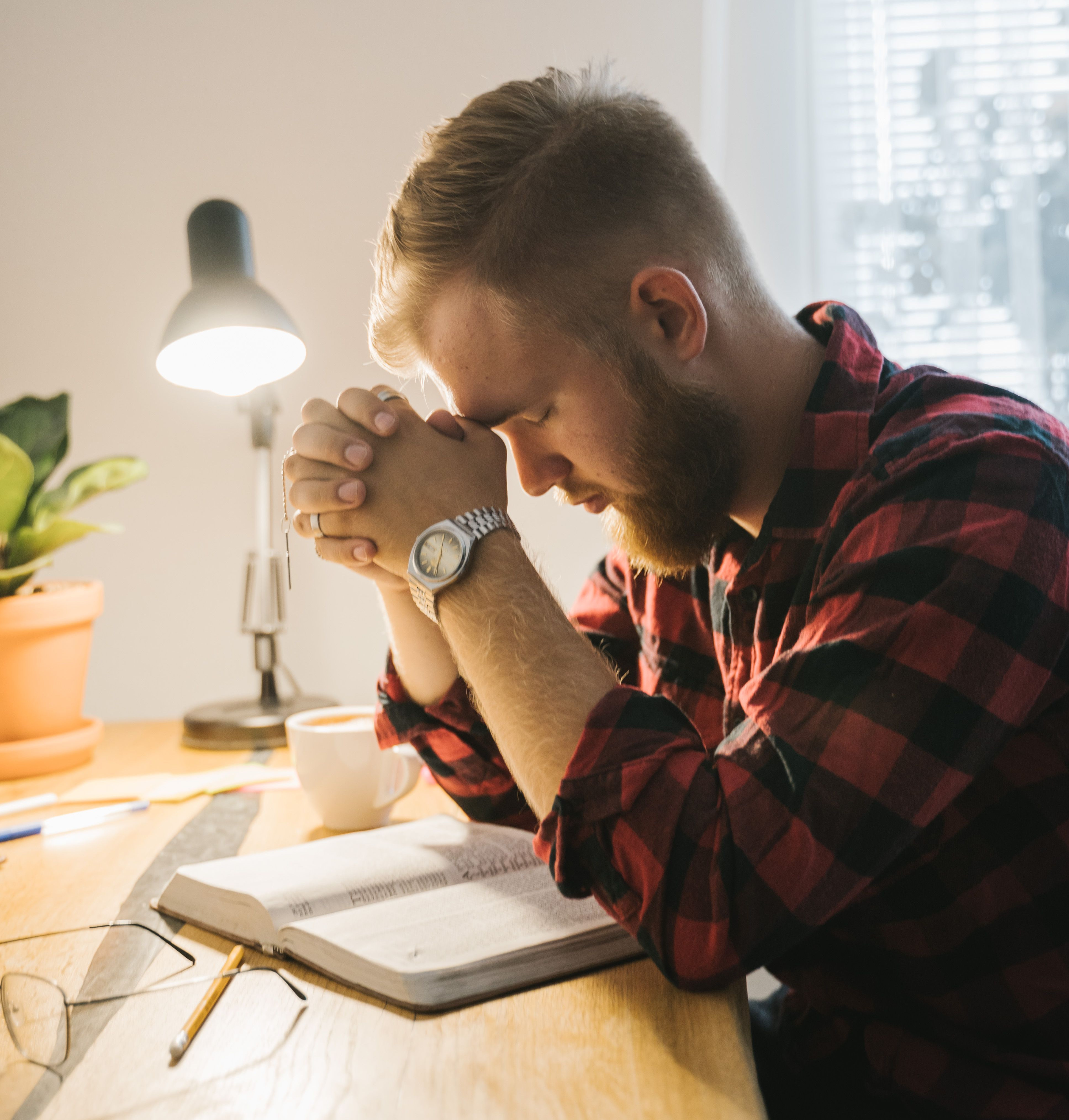 Bearded man in red plaid shirt prays at a desk with an open Bible, lamp, mug, and plant by a bright window.