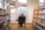 A man sits on a chair in the library, between two book shelves. He reads a newspaper facing the library window.