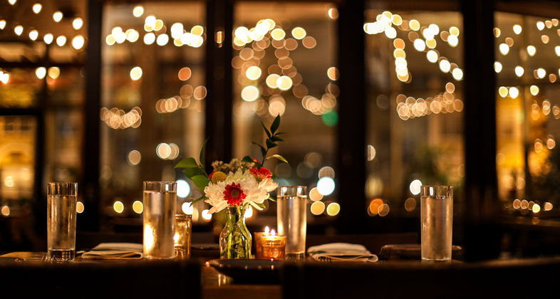 Restaurant dining room, dimly lit, set with water glasses and flowers, market lights in background