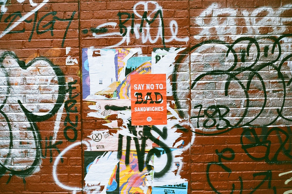 A wide photograph of a red brick wall covered in layered graffiti and torn posters. In the center, a bright orange flyer stands out with the bold text "SAY NO TO BAD SANDWICHES". It is pasted over a chaotic stack of weathered, ripped event posters. Surrounding the flyers, the brick wall is covered in multiple spray-painted graffiti pieces, including a large bubble letter outlined in black on the left, a looping white tag reading "SMILE" at the top with a heart above it, and dark green looping tags on the right side.