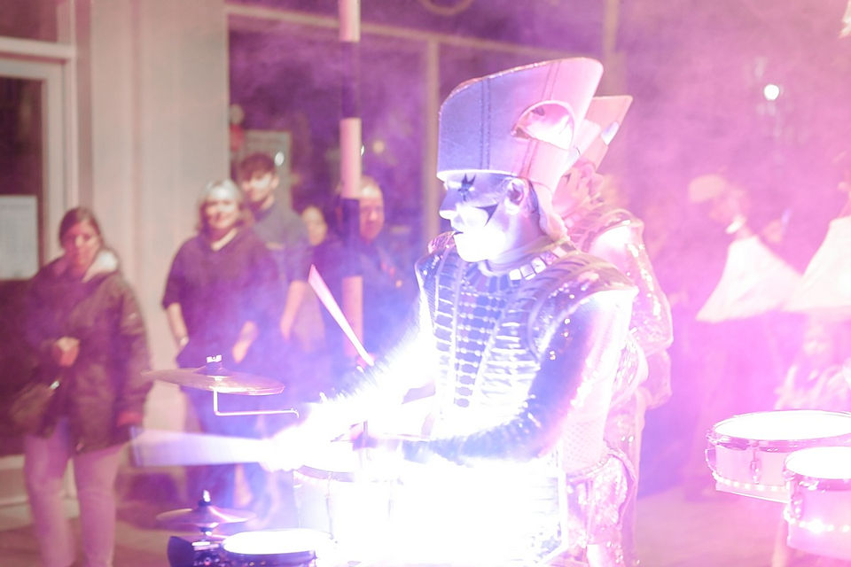 A medium shot capturing a Spark! drummer performing at night amidst thick pink theatrical smoke. The performer is shown from the waist up in a three-quarter profile facing left, wearing a futuristic silver cyber-armor vest, a tall structured headpiece, and geometric face makeup. Their drums and costume emit a brilliant, overexposed white and purple light that brightly illuminates their figure. In the blurred background on the left, several festival spectators stand on the sidewalk watching the procession.
