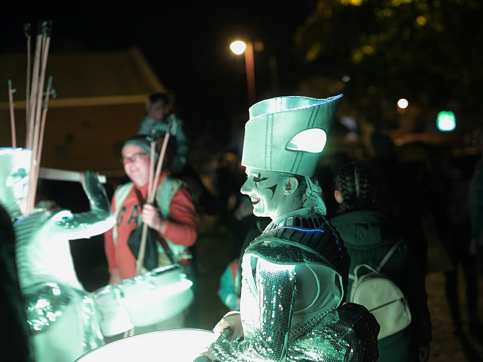 A medium night photograph showing a Spark! street performer smiling in profile, facing left as they stand among a festival crowd. The performer is brightly illuminated by an internal mint green LED light system, wearing a futuristic silver cyber-armor costume with a sequined sleeve and a tall, structured headpiece. To their left, another troupe member is seen playing an illuminated drum, while blurred festival attendees stand in the dark background beneath a warm orange street light.