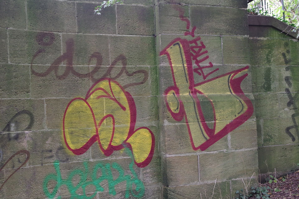 A photograph of a stone block wall covered in various styles of spray-painted graffiti. On the left side of the wall, the word "idea" is written in thin brown paint, positioned above a larger, bubbly yellow graffiti tag outlined in red. On the right side, an abstract geometric shape is painted in yellow with a thick magenta border, topped with smaller magenta scribbles. Faint green and black tags are also visible near the bottom and sides of the stone wall.