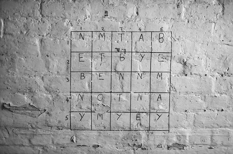A close-up photograph of a hand-drawn letter grid on a weathered brick wall.