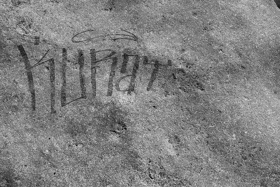 A black and white close-up photograph of graffiti scratched or carved into a rough, textured concrete surface. The word "KUNT" is faintly etched into the stone in a blocky, stylized script. A thin, looping line is drawn above the first few letters. The concrete background is heavily pitted, uneven, and covered in fine grains and dark smudges.