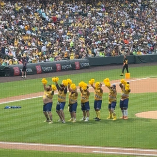 Seven Savannah Banana "Dad Bod Cheerleading Squad" ā the Man-Nanas ā are in a line in front of the pitcher's mound. They are holding yellow pom-poms above their heads.