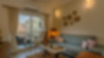 Second home interior design in Goa. 
Cozy living room with a teal sofa, vibrant cushions, and warm lighting. Wall decor, open book on table. Balcony view with greenery.