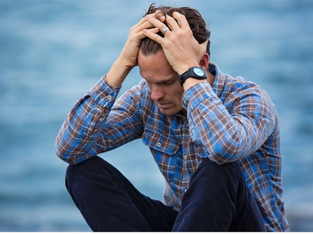 A man in a blue plaid shirt sits with his head in his hands, looking down with an overthinking expression. The background is a blurry blue, suggesting water or sky.