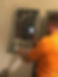 A technician in an orange shirt checks a wall-mounted furnace, using a handheld device to inspect settings or readings
