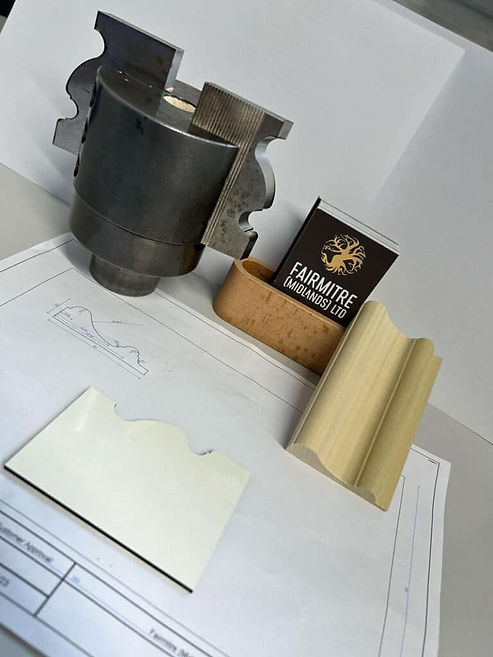 A metal cutting tool, a piece of wooden moulding, and a technical drawing are on a table