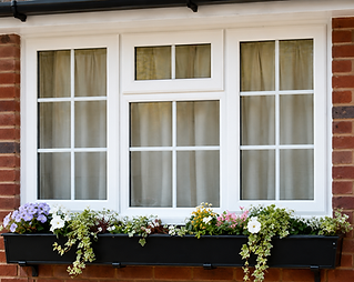 A white-framed window with beige curtains overlooks a windowsill adorned with lush green plants and colourful flowers, set against a brick wall