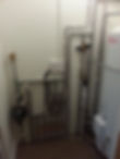 The corner of a room with a boiler and many pipes fitted on a wall