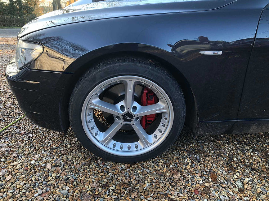 Close-up of the front wheel of a black car featuring a silver alloy wheel with red brake callipers on a gravel surface