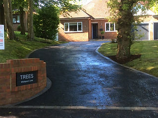 A brick house with a tarmac driveway, surrounded by lush greenery