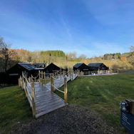 Wooden boardwalk or pathway leading towards a group of dark wooden holiday cabins on a grassy field