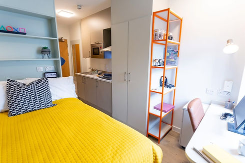 Compact, modern room with a vibrant yellow bedspread, orange shelving unit, and a small kitchenette