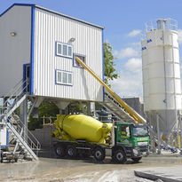 A green truck with a yellow cement mixer positioned under a cement plant structure