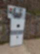 A white energy storage unit is mounted against a stone and wire wall with a gravel path