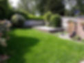 A back garden with green grass, trimmed bushes, and a wooden deck