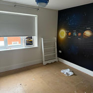 A room with solar system wallpaper on one wall, a window with a blind, and wooden flooring