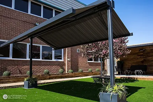 A modern patio awning shading a manicured lawn with potted plants and a brick wall with a large window