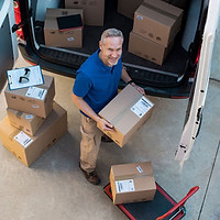 A person in a blue shirt is unloading boxes from a delivery van with packages nearby