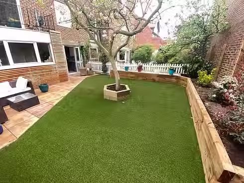 A vibrant garden with artificial grass, a tree in a wooden planter, surrounded by blooming plants