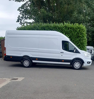 A white delivery van is parked on a paved surface in front of dense greenery
