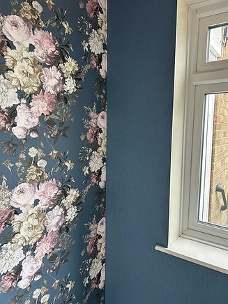 Floral wallpaper with pink and white blooms against a dark blue background contrasts a plain blue wall next to a window