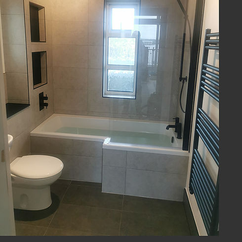 Modern bathroom with gray tiled walls, an L-shaped bathtub, glass shower screen, white toilet, window, and dark towel rail.