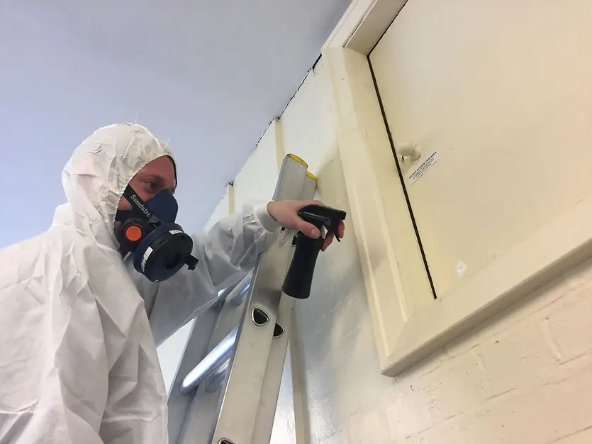A person in a protective suit sprays a cleaning solution on a wall during asbestos removal