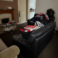 A cluttered living room with two sofas piled high with clothes and items