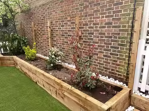 A wooden planter with several shrubs, bordered by white pebbles, set against a brick wall in a garden area
