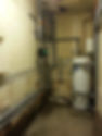 A water heater and pipes in a room with dampness on the wall